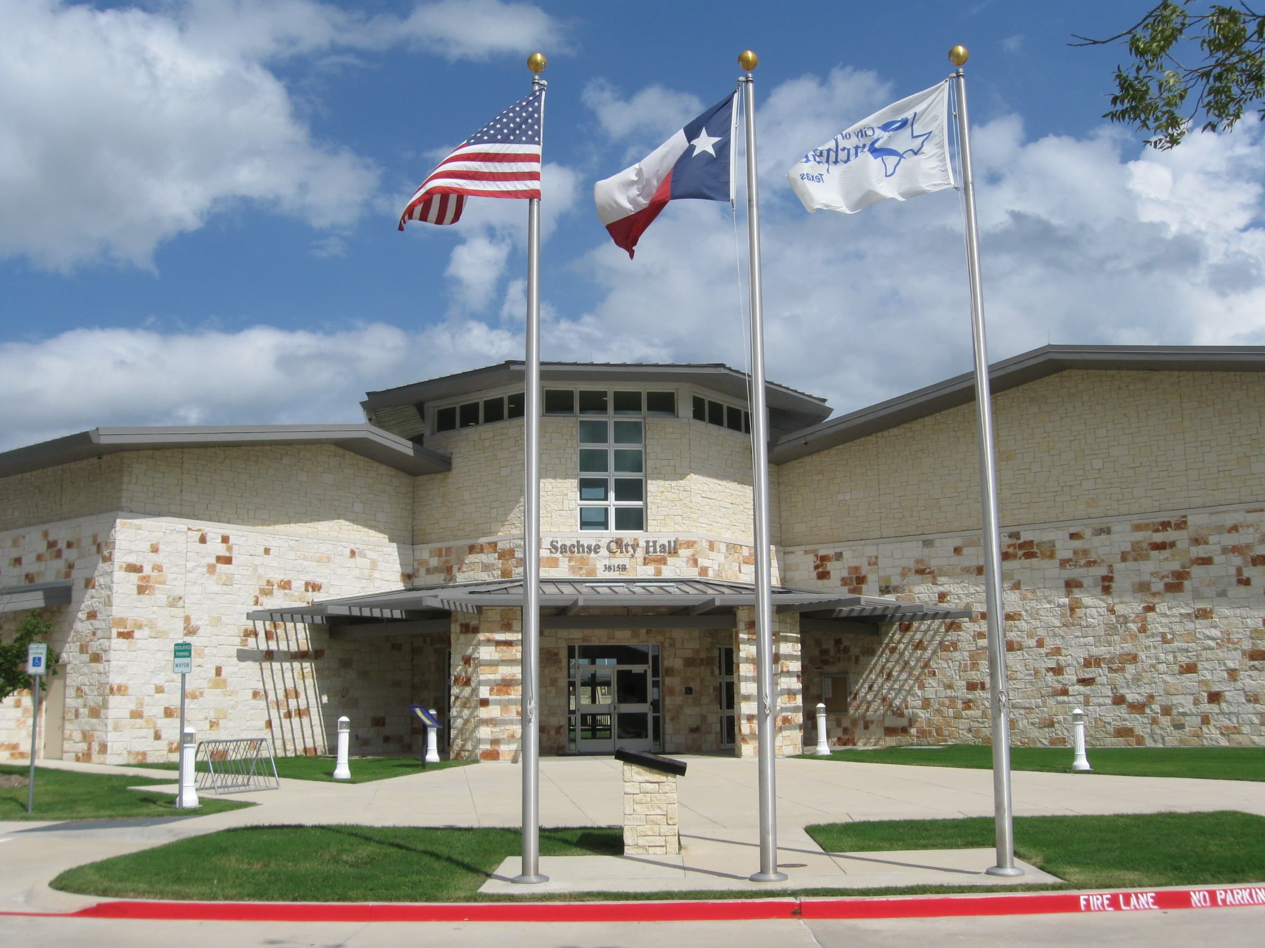 Edificio cívico de la alcaldía de Sachse, Texas, recubierto de piedra, con las banderas de Estados Unidos, Texas y la ciudad al frente